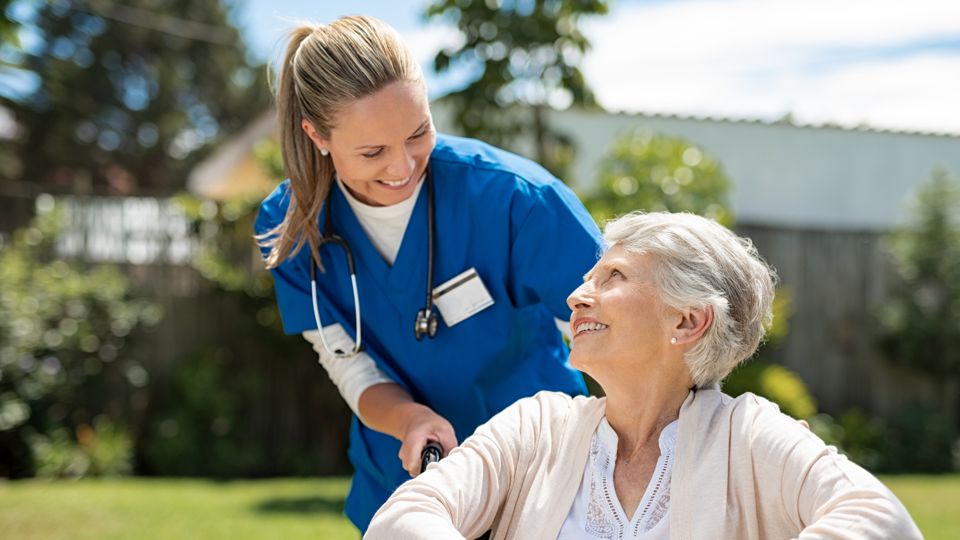 A healthcare professional and care home resident smile happily at each other while enjoying their time in the facility's garden